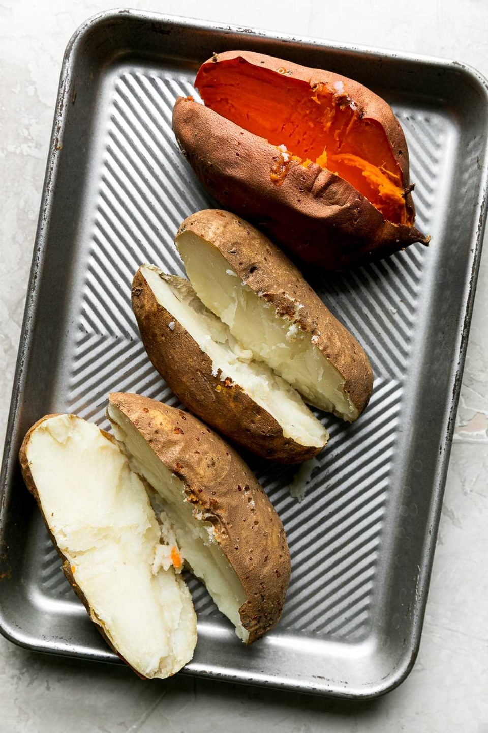 Two baked russet potatoes and one baked sweet potato that have all been cut in half lengthwise rest on a small aluminum cutting board. The cutting board rests on a creamy white plaster textured surface.