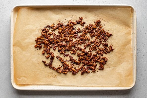 Roasted Maple Spiced Walnuts rest atop a piece of brown parchment paper that lines a cream colored baking sheet. The baking sheet sits atop a creamy white textured surface.