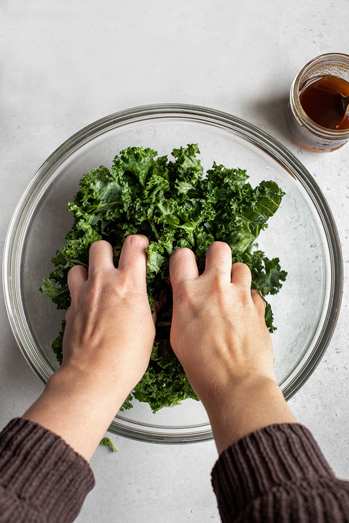 A clear glass mixing bowl filled with shredded kale and drizzled with cider-cinnamon vinaigrette is being massaged by a pair of woman's hands to help work the vinaigrette into the kale. A small mason jar filled with additional cider-cinnamon dressing rests alongside the mixing bowl and the bowl and mason jar sit atop a creamy white textured surface.