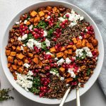 Thanksgiving salad in a large white serving bowl sits atop a white textured surface. A blue and white linen napkin, a few sprigs of fresh thyme, and a few pieces of pomegranate and loose pomegranate arils rests alongside the serving bowl. Two serving spoons rest inside the bowl.