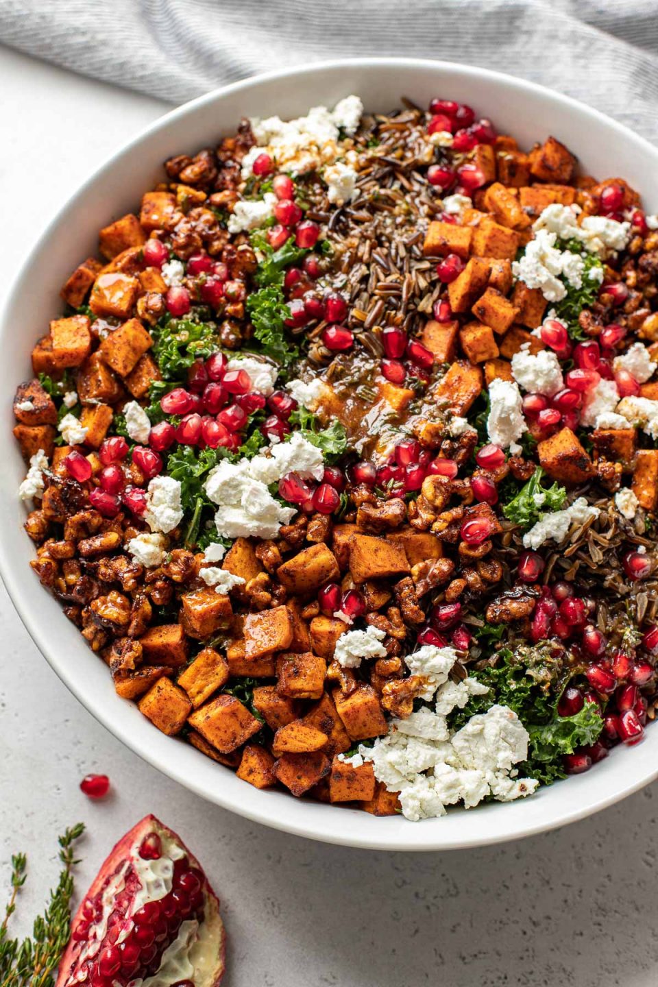 Thanksgiving salad in a large white serving bowl sits atop a white textured surface. A blue and white striped linen napkin, a few sprigs of fresh thyme, and pieces of pomegranate with loose pomegranate arils rests alongside the serving bowl.
