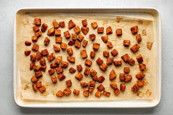Roasted Maple Spiced Sweet Potatoes rest atop a piece of brown parchment paper that lines a cream colored baking sheet. The baking sheet sits atop a creamy white textured surface.