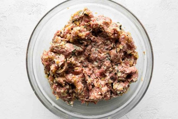 A large glass mixing bowl filled with mixed ground beef, ground pork, and a seasoned ricotta panko breadcrumb mixture sits atop a creamy white cement surface.