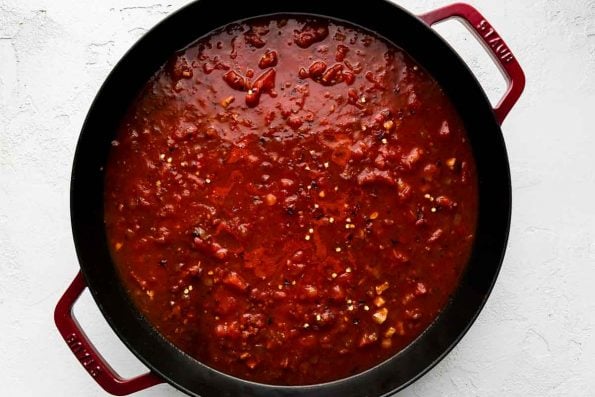 A simple tomato sauce simmers in a cherry red Staub 13-inch Double Handle Fry Pan. The fry pan rests atop a creamy white cement surface.