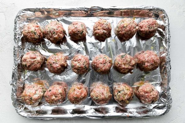 18 uncooked golf ball sized meatballs are arranged atop an aluminum foil lined baking sheet. The baking sheet rests atop a creamy white cement surface.