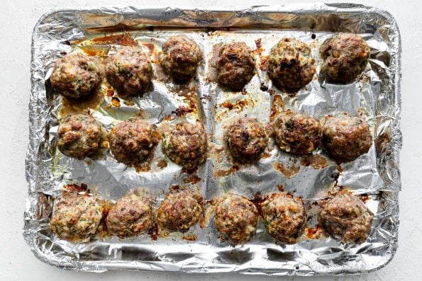 18 baked golf ball sized meatballs are arranged atop an aluminum foil lined baking sheet. The baking sheet rests atop a creamy white cement surface. A blue and white striped linen napkin rests alongside the baking sheet.