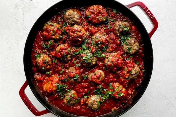 Best-Ever Ricotta Meatballs simmer in a homemade simple red sauce in a cherry red Staub 13-inch Double Handle Fry Pan. The meatballs have been garnished with freshly chopped basil and parsley and the fry pan sits atop a creamy white cement surface.