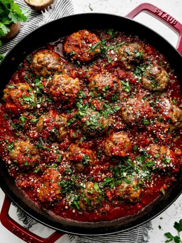 Best-Ever Ricotta Meatballs simmer in a homemade simple red sauce in a cherry red Staub 13-inch Double Handle Fry Pan. The meatballs have been garnished with freshly chopped basil, parsley, and parmesan cheese and the fry pan sits atop a creamy white cement surface. A blue and white striped linen napkin has been placed underneath the fry pan. A small bouquet of fresh parsley in a wooden container and a small wooden pinch bowl filled with parmesan cheese sit atop the linen napkin, alongside of the fry pan.