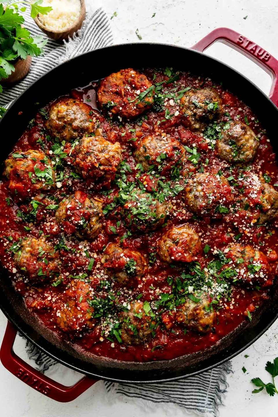 Best-Ever Ricotta Meatballs simmer in a homemade simple red sauce in a cherry red Staub 13-inch Double Handle Fry Pan. The meatballs have been garnished with freshly chopped basil, parsley, and parmesan cheese and the fry pan sits atop a creamy white cement surface. A blue and white striped linen napkin has been placed underneath the fry pan. A small bouquet of fresh parsley in a wooden container and a small wooden pinch bowl filled with parmesan cheese sit atop the linen napkin, alongside of the fry pan.