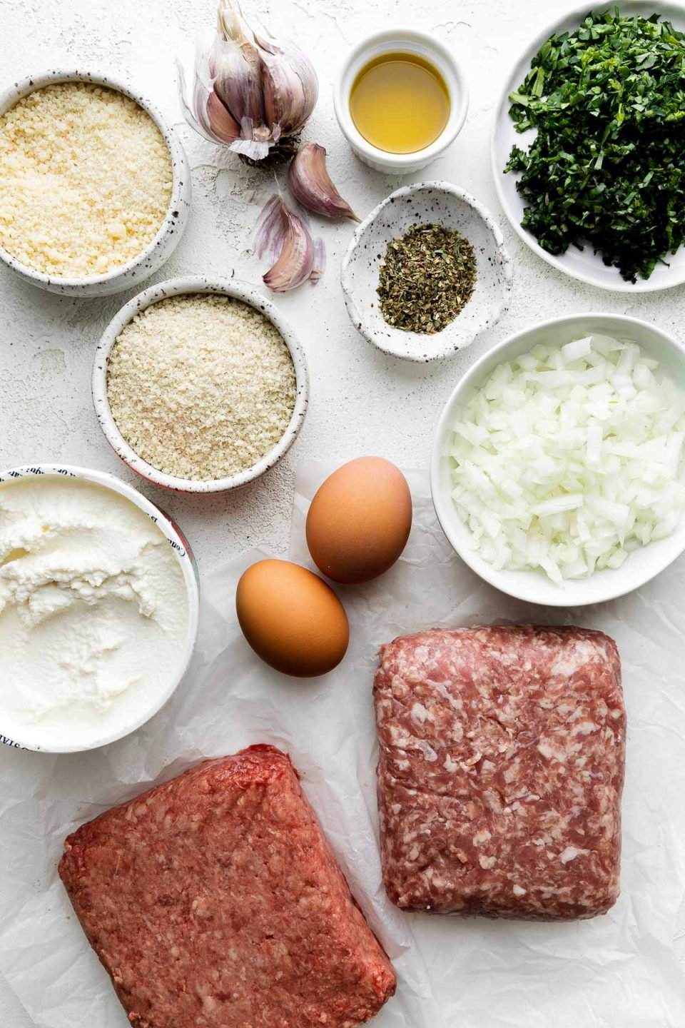 Best-Ever Ricotta Meatballs ingredients arranged on a creamy cement surface - ground beef and pork atop a sheet of crinkled wax paper, olive oil, yellow onion, garlic, whole milk ricotta, panko breadcrumbs, eggs, grated parmesan, fresh parsley, fresh basil, Italian seasoning.