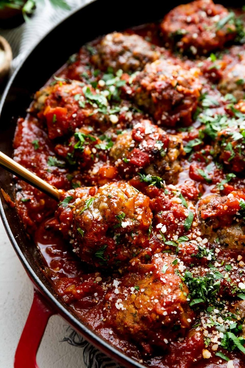 A side angle shot of Best-Ever Italian Meatballs inside of a homemade simple red sauce in a cherry red Staub 13-inch Double Handle Fry Pan. The meatballs have been garnished with freshly chopped basil, parsley, and parmesan cheese and the fry pan sits atop a creamy white cement surface. A blue and white striped linen napkin has been placed underneath the fry pan. A small bouquet of fresh parsley in a wooden container and a small wooden pinch bowl filled with parmesan cheese sit alongside of the fry pan. A gold spoon rests inside of the pan lifting up a single meatball.