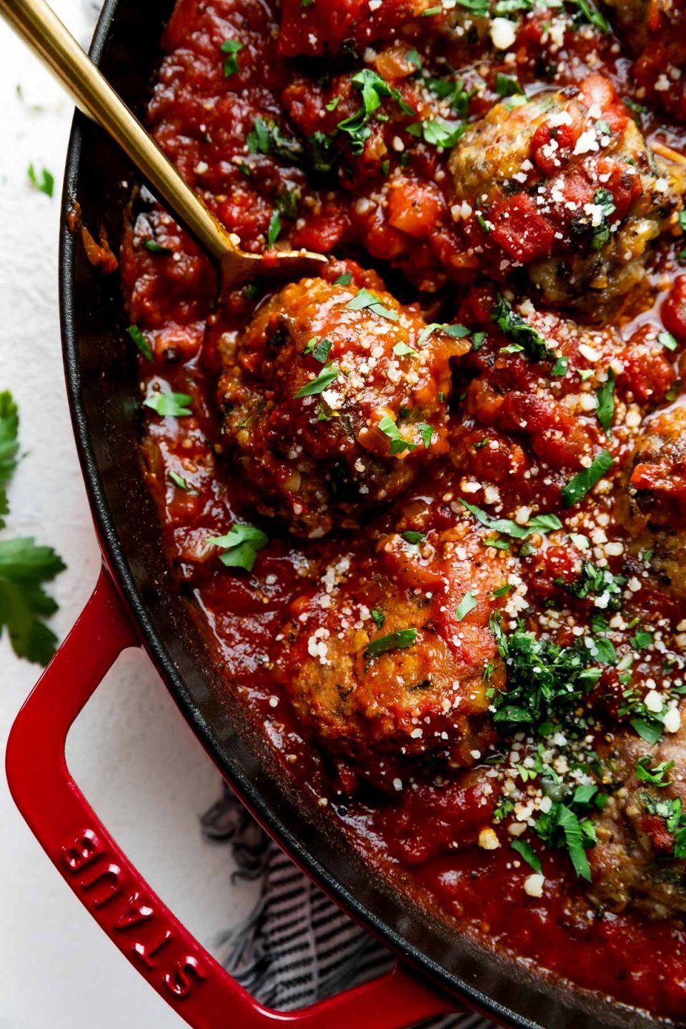 A side angle shot of Best-Ever Italian Meatballs inside of a homemade simple red sauce in a cherry red Staub 13-inch Double Handle Fry Pan. The meatballs have been garnished with freshly chopped basil, parsley, and parmesan cheese and the fry pan sits atop a creamy white cement surface. A blue and white striped linen napkin has been placed underneath the fry pan. A few loose fresh parsley leaves rest alongside of the fry pan and a gold spoon rests inside of the pan lifting up a single meatball.