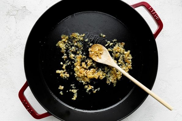 Chopped yellow onion and garlic softens in olive oil inside of a cherry red 13-inch Staub Double Handle Fry Pan. The fry pan sits atop a creamy white cement surface with a wooden spoon resting inside of the pan for mixing.