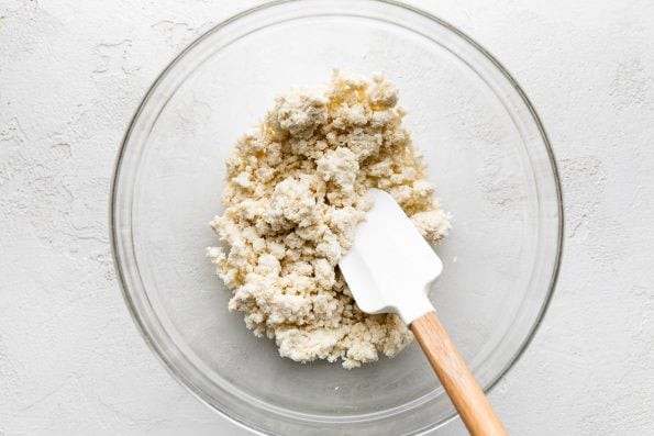 A mixture of panko breadcrumbs and whole milk ricotta cheese are stirred together and rest inside of a large glass mixing bowl. A wooden spatula with a white rubber head rest inside of the bowl used for mixing.