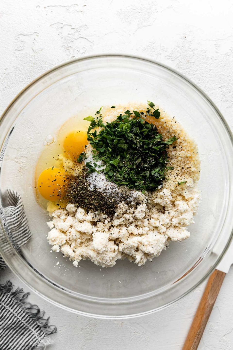 A large glass mixing bowl filled with ricotta soaked panko breadcrumbs, eggs, grated parmesan cheese, softened onion and garlic, freshly chopped parsley and basil, and Italian seasoning that will become a seasoned breadcrumb mixture for Italian ricotta meatballs. A wooden spatula and a white and blue striped linen napkin rest alongside the mixing bowl and the bowl sits atop a creamy white cement surface.