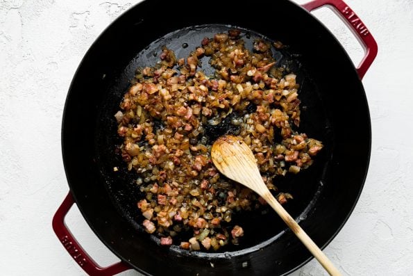 Pancetta and yellow onions render and soften inside of a cherry red Staub 13-inch Double Handle Fry Pan. A wooden spoon rests inside of the fry pan and the fry pan sits atop a creamy white cement surface.