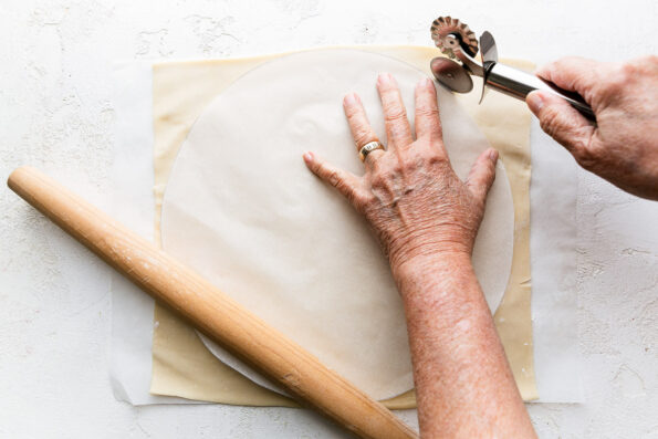 Puff pastry sheets rest atop a single sheet of parchment paper. An 11-inch circular template cut from parchment paper rests on top of the puff pastry sheet. A woman's hands work to hold the template in place with one hand while holding a pastry cutter in the other hand carefully cutting the puff pastry into an 11-inch circle. The pastry sheet and parchment paper rests atop a creamy white textured surface and a tapered wooden rolling pin rests alongside the puff pastry sheet.