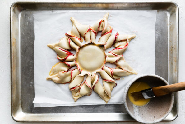 A puff pastry star filled with spiced raspberry jam and cream cheese filling sits atop a baking sheet lined with parchment paper. A ceramic bowl filled with egg wash sits alongside the star on the baking sheet. A pastry brush sits inside of the bowl and the baking sheet sits atop a creamy white textured surface.