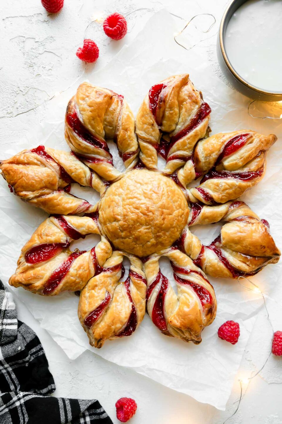 A baked Christmas Kringle puff pastry star sits atop a couple sheets of crinkled parchment paper. The parchment paper sits atop a creamy white textured surface. A small bowl filled with icing rests alongside the puff pastry star with loose fresh raspberries, twinkling lights, and a black and white plaid linen napkin surround the puff pastry star.