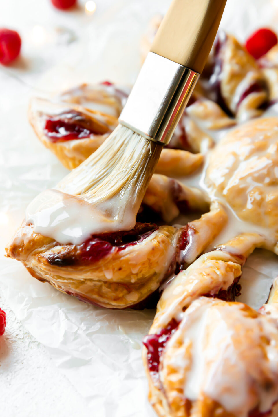 A side angle shot of baked Christmas Kringle puff pastry star with icing. A pastry brush brushes additional icing on the puff pastry star. The pastry sits atop a piece of parchment paper. Loose raspberries surround the pastry.