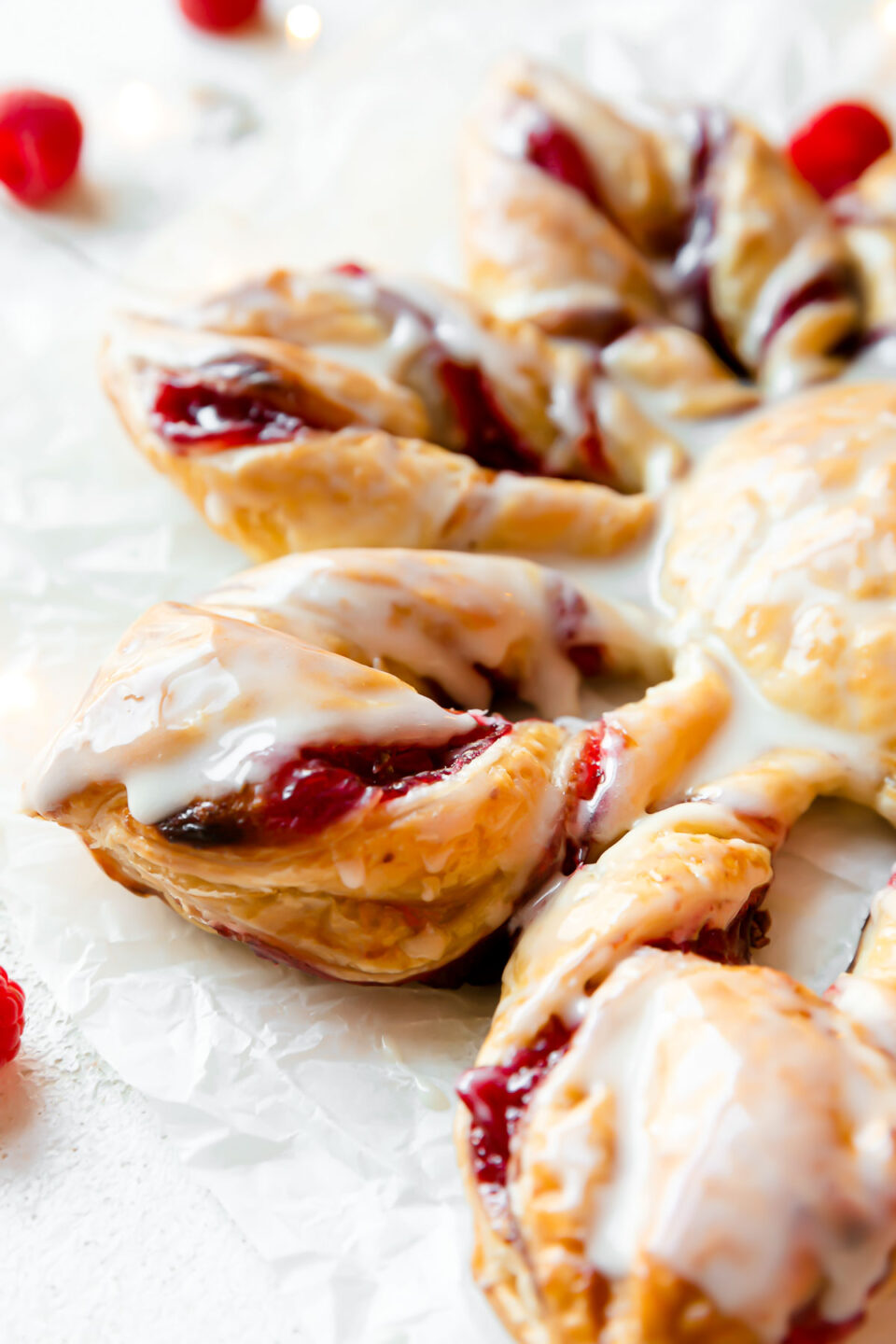 A side angle shot of baked Christmas Kringle puff pastry star with icing. The pastry sits atop a piece of parchment paper. Loose raspberries surround the pastry.