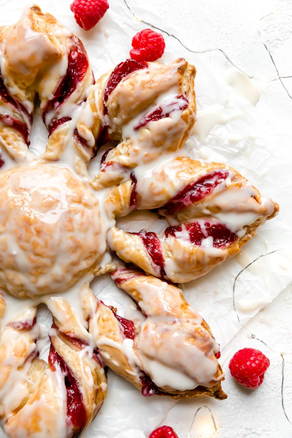 A Christmas Kringle Puff Pastry Star sits atop a piece of parchment paper. A string of twinkling lights and fresh raspberries surround the pastry. The pastry sits atop a creamy white textured surface.