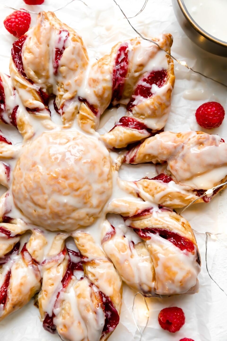 A Christmas Kringle Puff Pastry Star sits atop a piece of parchment paper. A string of twinkling lights, fresh raspberries, and a bowl of icing surround the pastry. The pastry sits atop a creamy white textured surface.