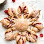 A Christmas Kringle Puff Pastry Star sits atop a piece of parchment paper. A string of twinkling lights, fresh raspberries, and a bowl of icing surround the pastry. The pastry sits atop a creamy white textured surface.