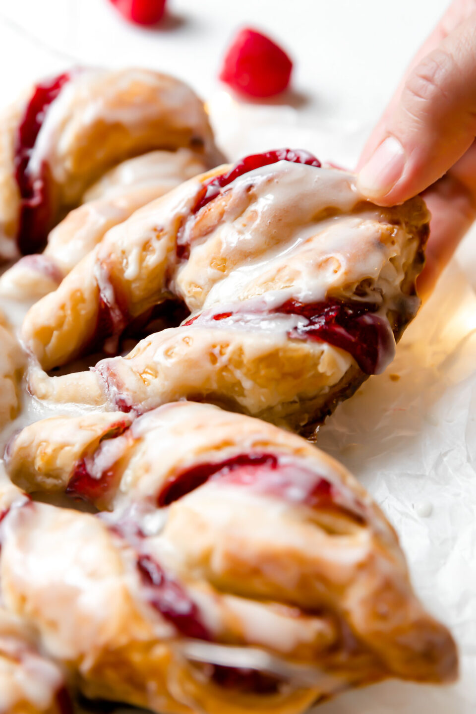 A side angle shot of Christmas Kringle Puff Pastry Star sits atop a piece of parchment paper. Loose fresh raspberries rest alongside the pastry in the background and the pastry sits atop a creamy white textured surface. A woman's hand reaches for a section of the puff pastry star.