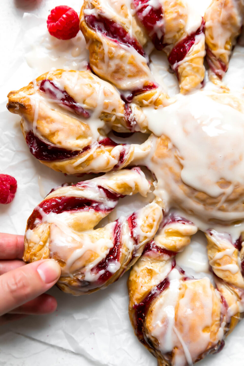 An overhead shot of Christmas Kringle Puff Pastry Star sits atop a piece of parchment paper. Loose fresh raspberries rest alongside the pastry in the background and the pastry sits atop a creamy white textured surface. A woman's hand reaches for a section of the puff pastry star.