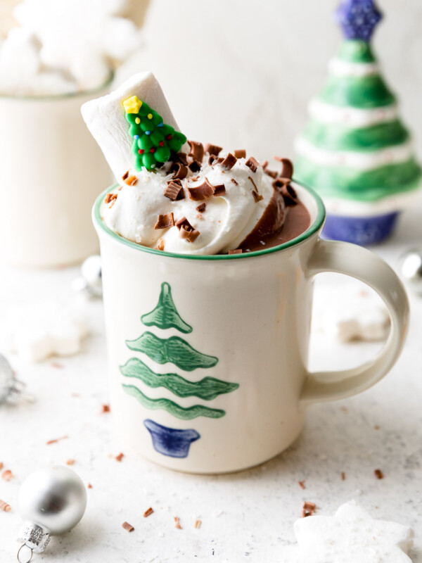 A white mug decorated with a Christmas tree is filled with Christmas Hot Chocolate, topped with whipped cream, & garnished with chocolate shavings & a Christmas tree marshmallow. The mug sits atop a white surface alongside marshmallows, ornaments, & small Christmas decorations.