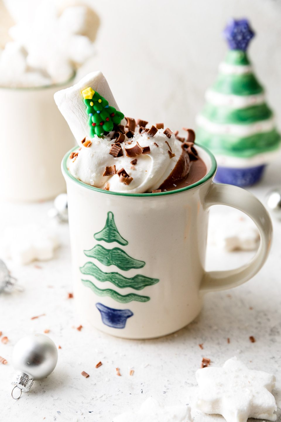 A white mug decorated with a Christmas tree is filled with Christmas Hot Chocolate, topped with whipped cream, & garnished with chocolate shavings & a Christmas tree marshmallow. The mug sits atop a white surface alongside marshmallows, ornaments, & small Christmas decorations.