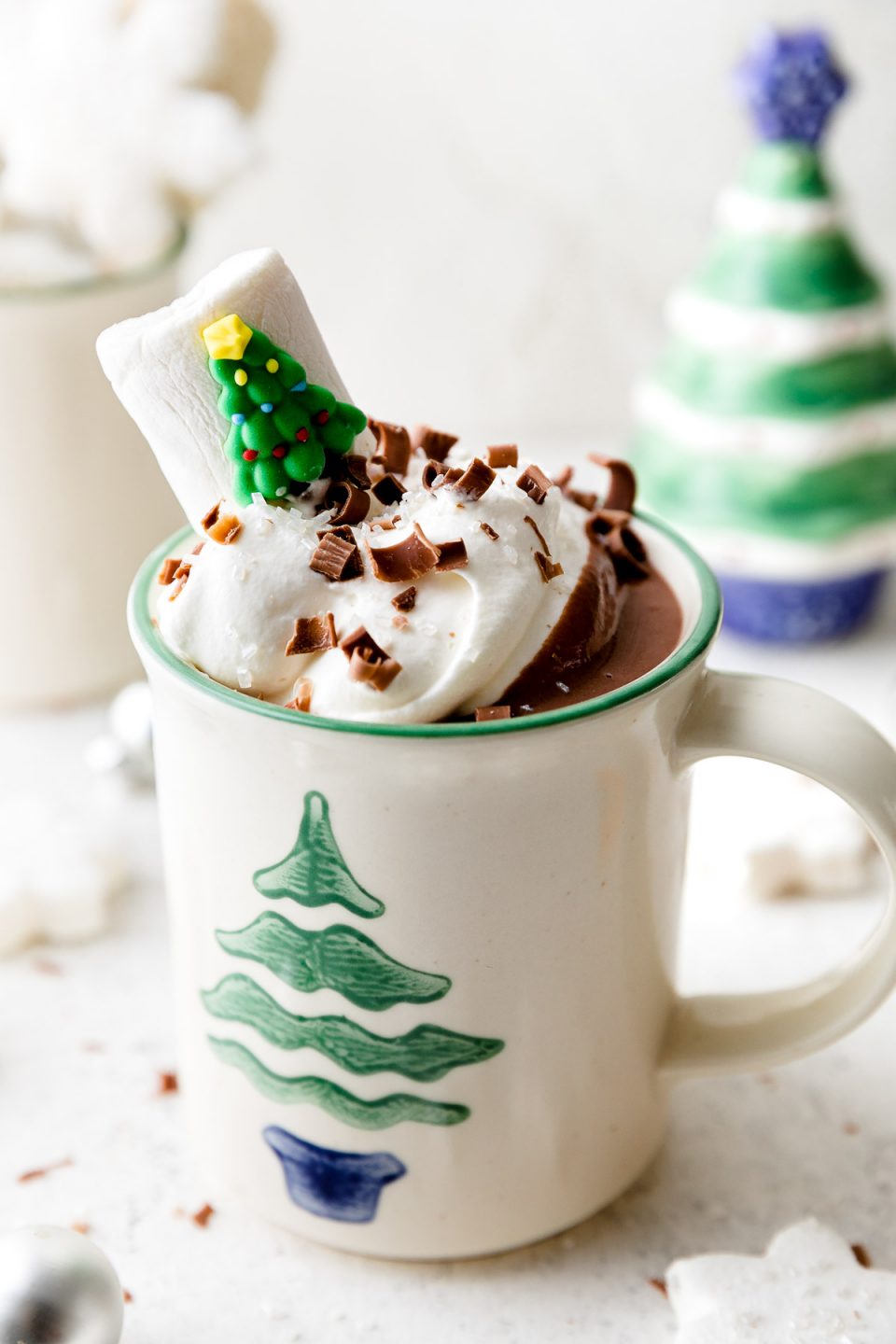 A white mug decorated with a Christmas tree is filled with Christmas Hot Chocolate, topped with whipped cream, & garnished with chocolate shavings & a Christmas tree marshmallow. The mug sits atop a white surface alongside marshmallows, ornaments, & small Christmas decorations.