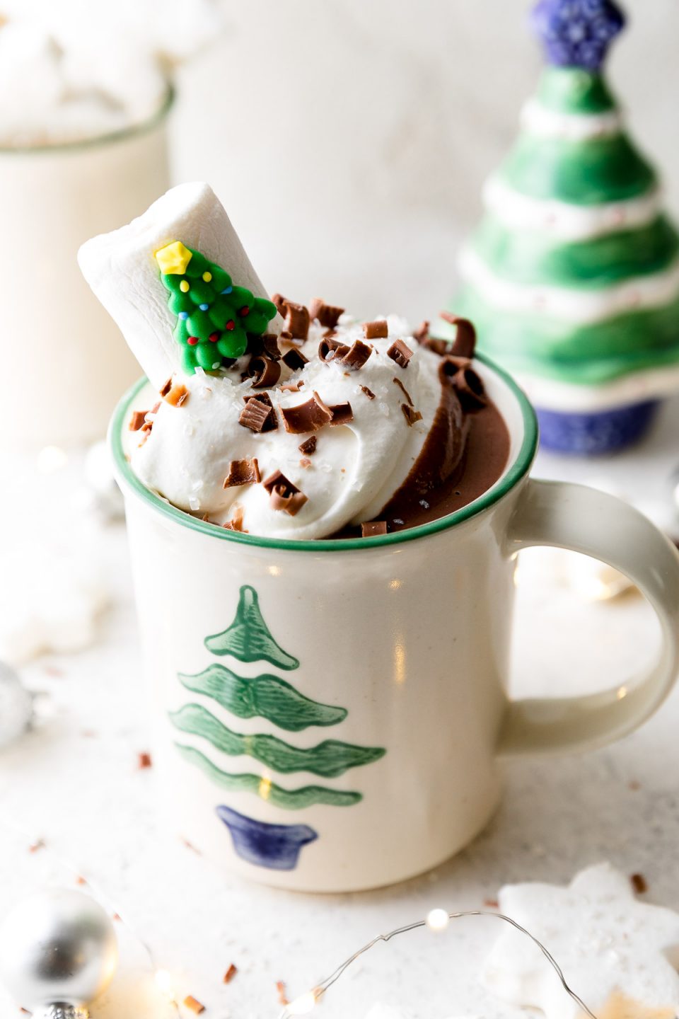A white mug decorated with a Christmas tree is filled with Christmas Morning Cocoa, topped with whipped cream, & garnished with chocolate shavings & a Christmas tree marshmallow. The mug sits atop a white surface alongside marshmallows, ornaments, & small Christmas decorations.