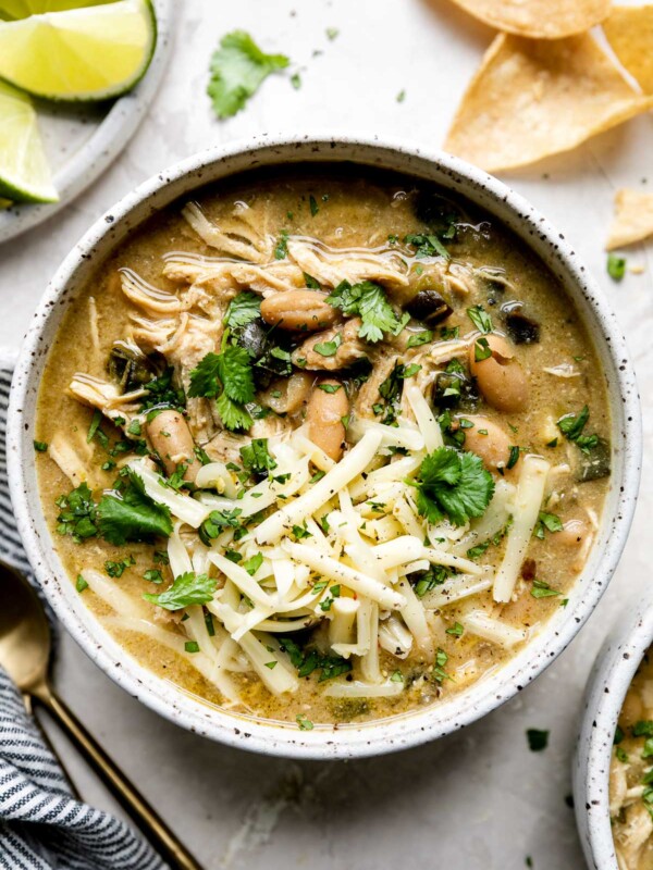 Creamy White Chili served in a gray speckled ceramic bowl sits atop a white textured surface. The chili is garnished with chopped cilantro, cheese, and ground black pepper. A second bowl of chili, a plate of lime wedges, loose tortilla chips, cilantro leaves, two gold spoons, and a blue and white striped linen napkin surround the bowl of chili.