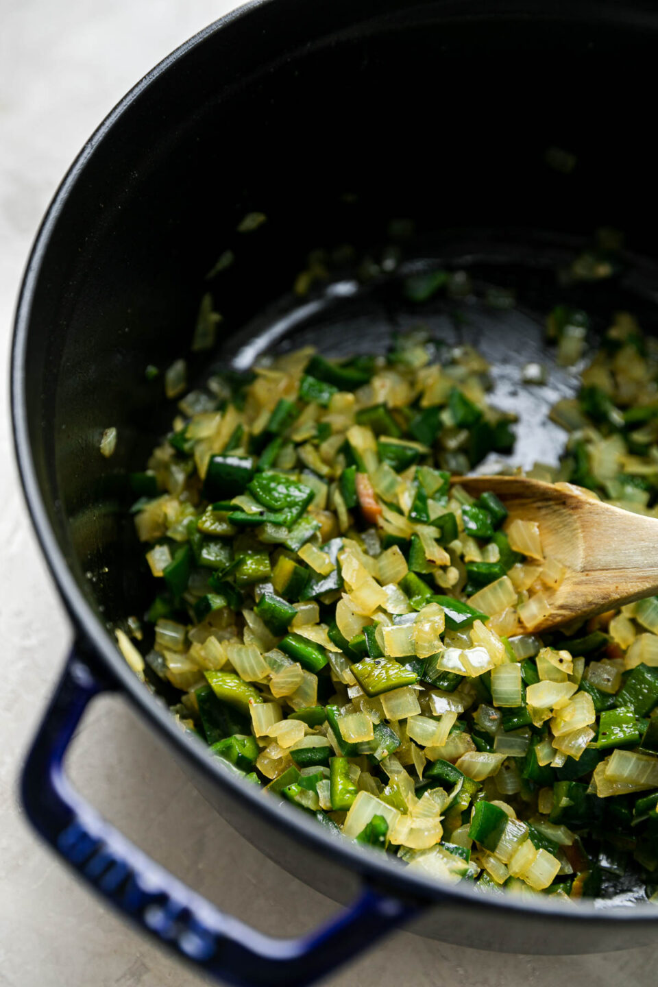 Diced yellow onion and poblano peppers soften inside of a blue Staub dutch oven. A wooden spoon rests inside of the dutch oven and the dutch oven sits atop a creamy white textured surface.