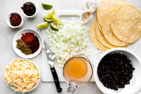 Crispy Black Bean Taco ingredients arranged on a creamy white textured surface: yellow onion, garlic, chipotle pepper, tomato paste, chili powder, ground cumin, smoked paprika, black beans, vegetable broth, lime, corn tortillas, Pepper Jack cheese.