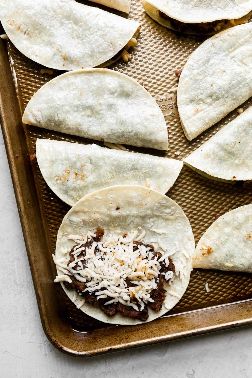 10 assembled black bean tacos arranged on a baking sheet. 9 of the tacos have been assembled with the tortillas folded, while one remains unfolded revealing the black bean mixture spread on one side of the tortillas with Pepper Jack cheese sprinkled over top. The baking sheet rests atop a creamy white textured surface.