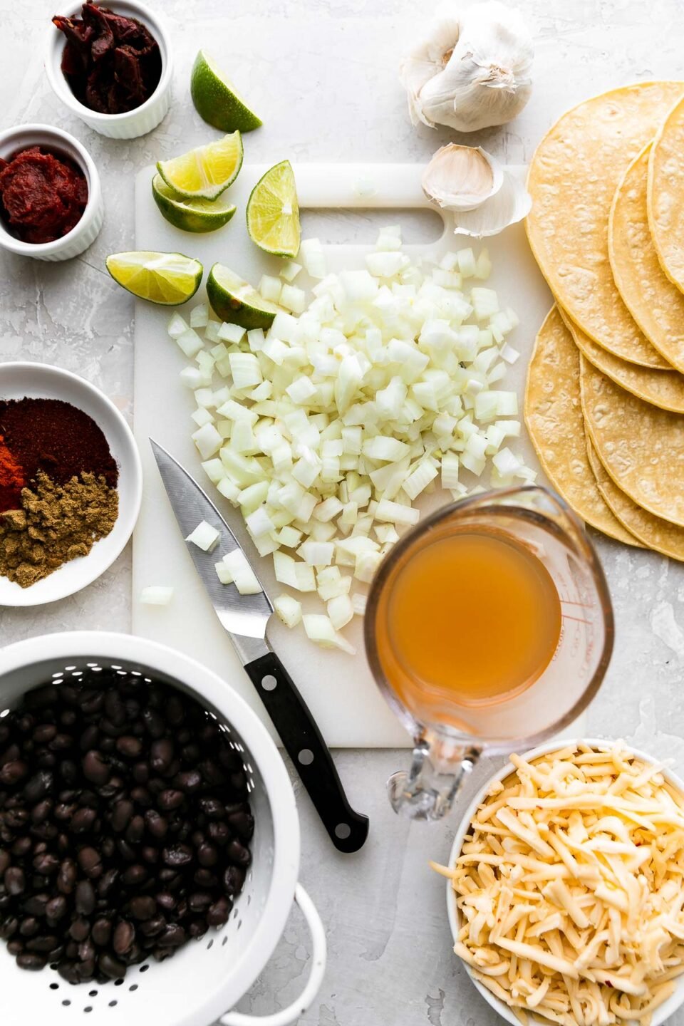 An overhead shot of ingredients arranged on a creamy white textured surface: yellow onion, garlic, chipotle pepper, tomato paste, chili powder, ground cumin, smoked paprika, black beans, vegetable broth, lime, corn tortillas, Pepper Jack cheese.