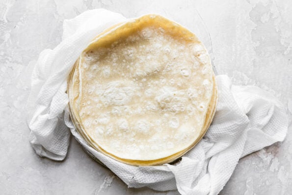 A stack of warmed corn tortillas sits atop damp paper towels. The paper towels sit atop a creamy white textured surface.