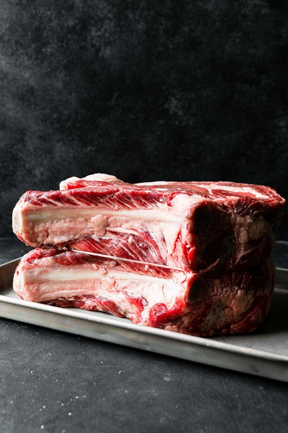 A side angle shot of a bone-in standing beef rib roast for prime rib turned on its side & sitting atop an aluminum baking sheet. The prime rib roast bones have been sliced off and then tied back on with kitchen twine. The baking sheet rests atop a dark textured surface with a dark textured background set behind it.