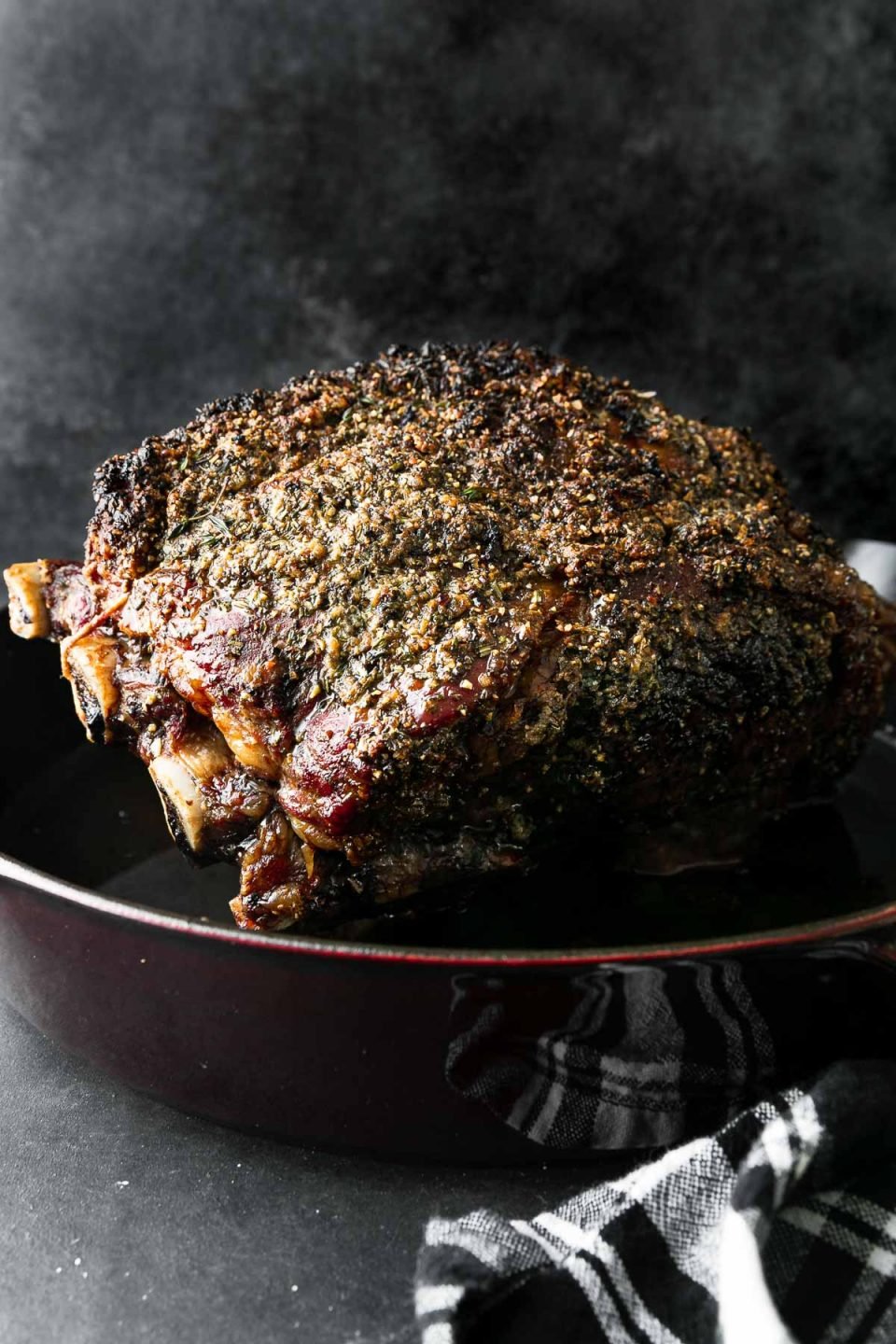 A side angle shot of roasted Prime Rib covered in a Garlic Herb Rub sits inside of a cherry red Staub cast iron Deep Skillet. The skillet sits atop a dark textured surface with a black and white plaid linen napkin resting alongside.