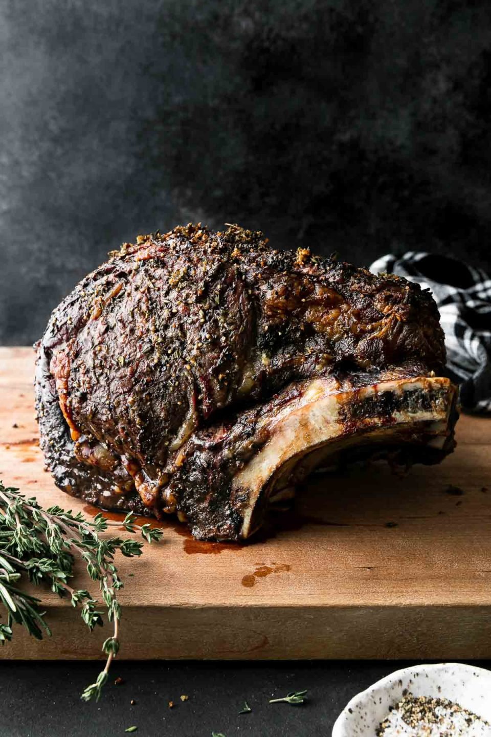 A side angle shot of roasted Prime Rib covered in a Garlic Herb Rub sitting atop a wooden cutting board. The cutting board rests atop a dark textured surface with a black and white plaid linen napkin resting in the background & fresh herbs, and a pinch bowl filled with kosher salt and black pepper in the foreground. The items are set against a dark textured background.
