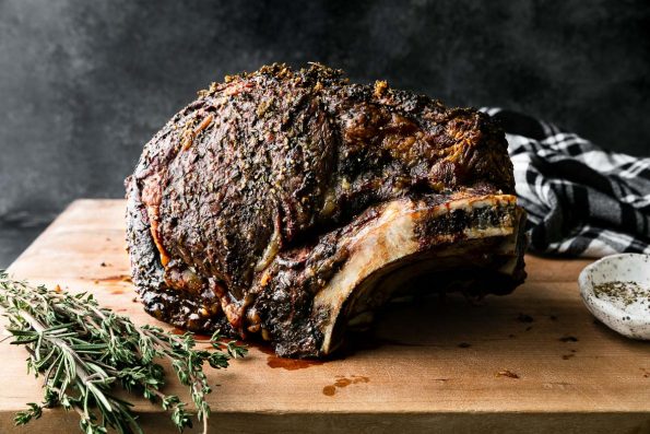 A side angle shot of roasted Prime Rib covered in a Garlic Herb Rub sitting atop a wooden cutting board. The cutting board rests atop a dark textured surface with a black and white plaid linen napkin resting in the background & fresh herbs, and a pinch bowl filled with kosher salt and black pepper surrounding the prime rib roast. The items are set against a dark textured background.