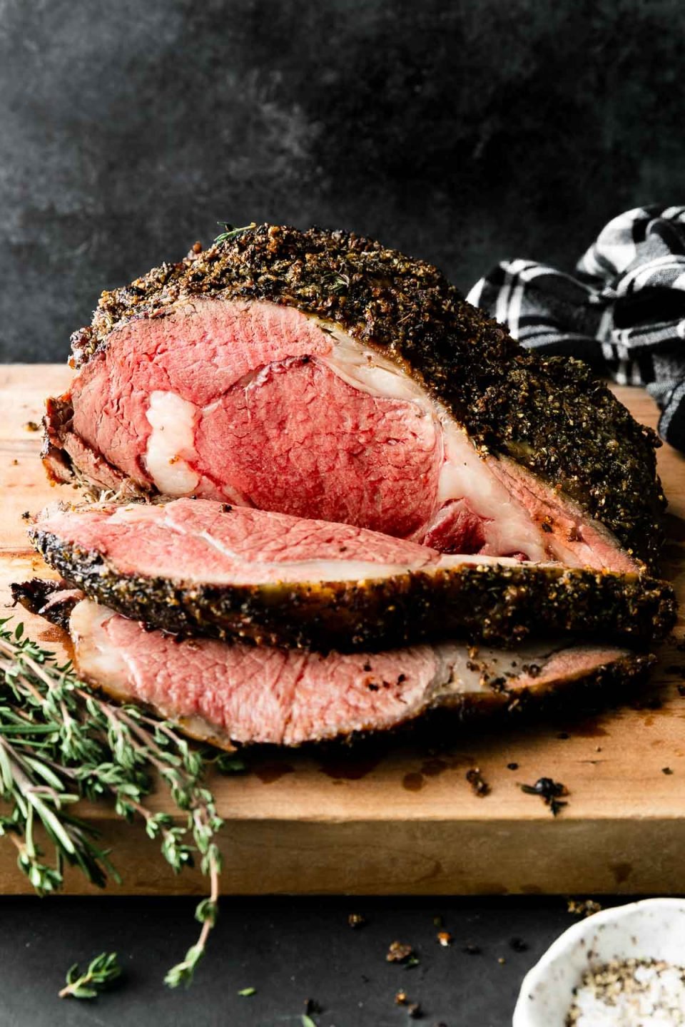 A side angle shot of roasted and partially carved medium-rare Prime Rib covered in a Garlic Herb Rub that sits atop a wooden cutting board. The cutting board rests atop a dark textured surface with a black and white plaid linen napkin resting in the background & fresh herbs, and a pinch bowl filled with kosher salt and black pepper surrounding the prime rib roast. The items are set against a dark textured background.