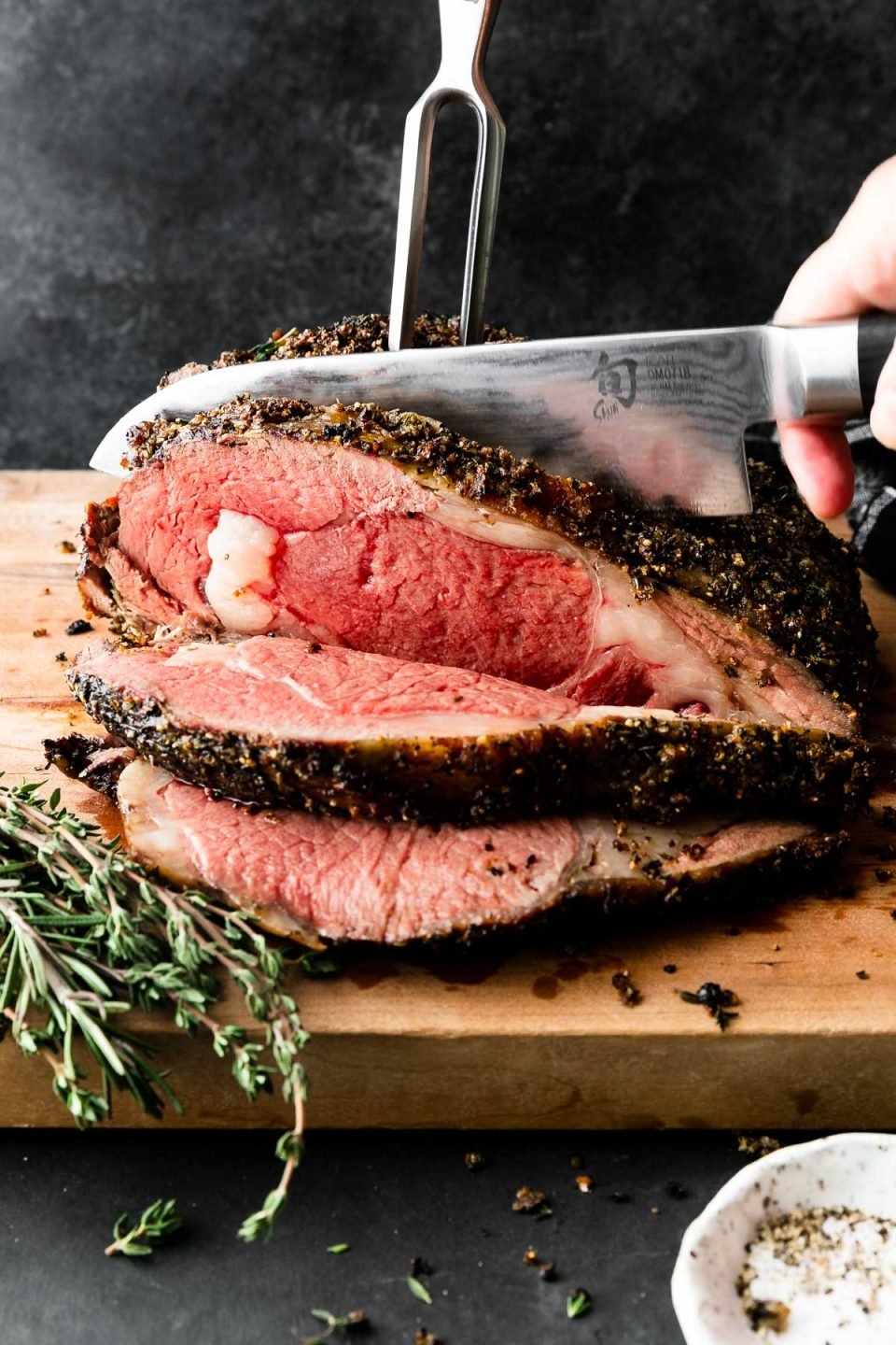 A side angle shot of roasted and partially carved medium-rare Prime Rib covered in a Garlic Herb Rub that sits atop a wooden cutting board. A woman's hand holds a sharp kitchen knife in one hand as it slices through the roast, while a carving fork stabilizes the prime rib roast behind the knife. The cutting board rests atop a dark textured surface with a black and white plaid linen napkin resting in the background & fresh herbs, and a pinch bowl filled with kosher salt and black pepper surrounding the prime rib roast. The items are set against a dark textured background.