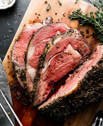 An overhead shot of roasted and partially carved medium-rare Prime Rib covered in a Garlic Herb Rub that sits atop a wooden cutting board. Fresh herbs, a sharp kitchen knife, and a carving fork surround the roast and rest alongside on the cutting board. The cutting board rests atop a dark textured surface with a black and white plaid linen napkin and a pinch bowl filled with kosher salt surrounding the cutting board.