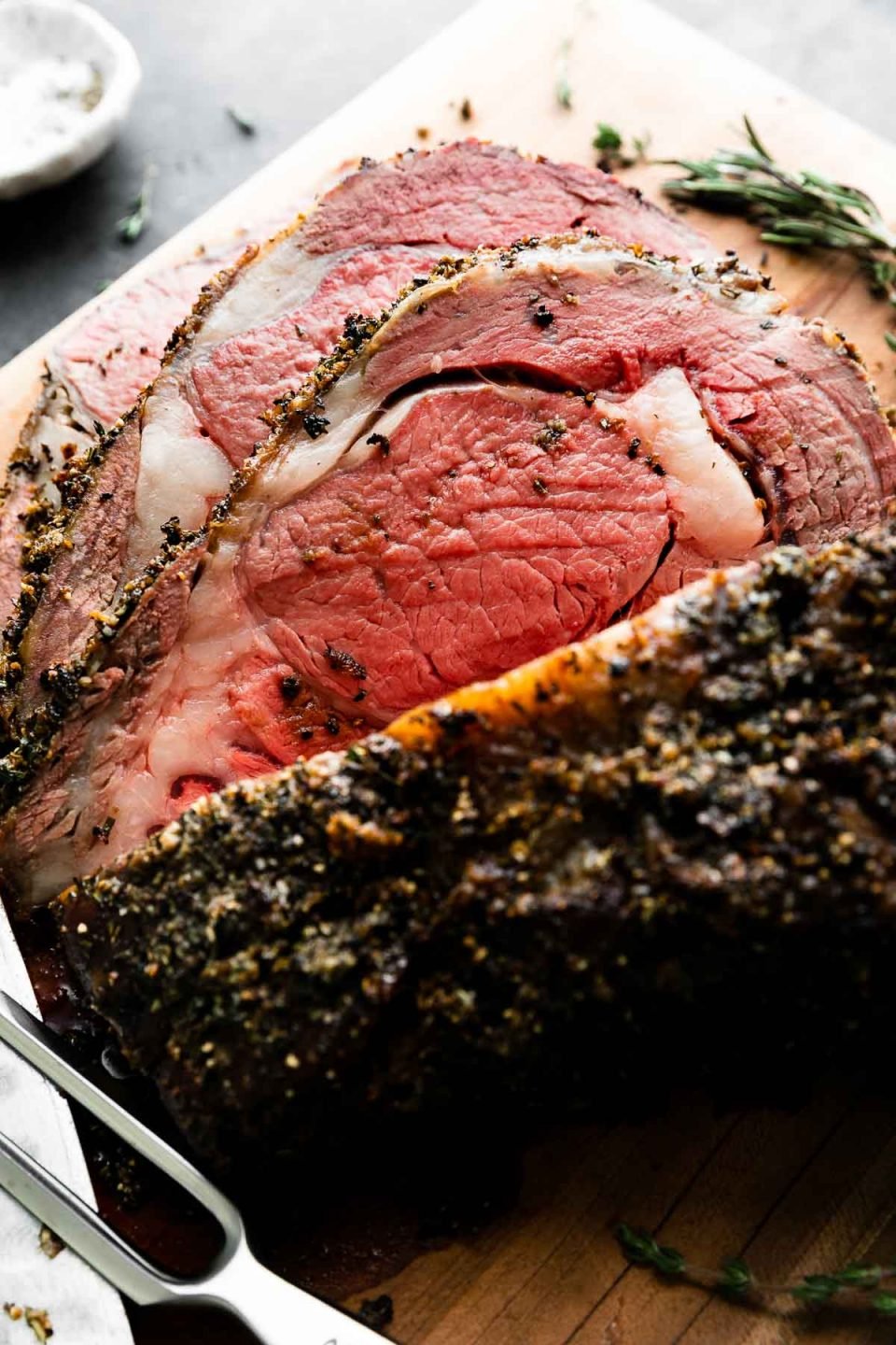 A side angle shot of roasted and partially carved medium-rare Prime Rib covered in a Garlic Herb Rub that sits atop a wooden cutting board. Fresh herbs, a sharp kitchen knife, and a carving fork surround the roast and rest alongside on the cutting board. The cutting board rests atop a dark textured surface with a black and white plaid linen napkin and a pinch bowl filled with kosher salt surrounding the cutting board.