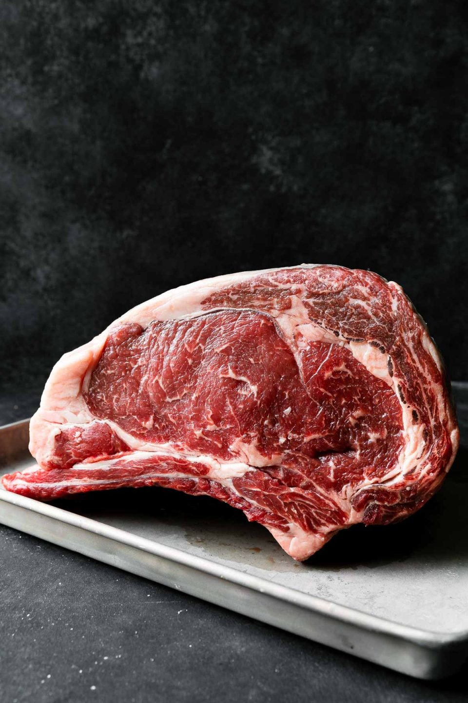 A side angle shot of a bone-in standing beef rib roast for prime rib sits atop an aluminum baking sheet. The baking sheet rests atop a dark textured surface with a dark textured background set behind it.