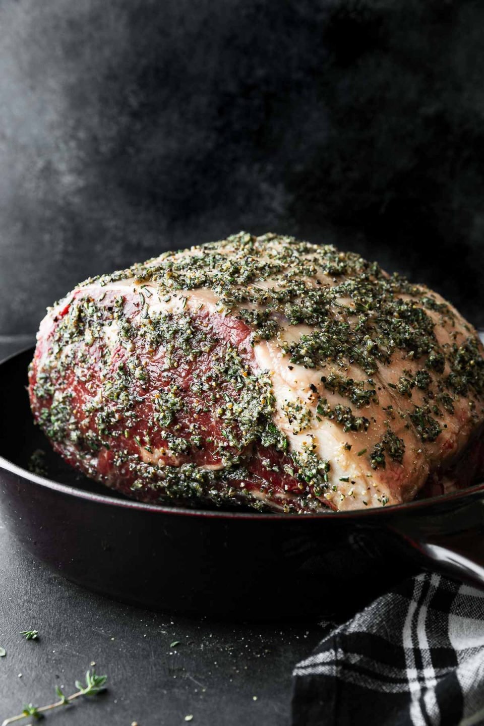 An uncooked bone-in standing prime rib roast seasoned with a garlic herb butter sits inside a red Staub Deep Skillet. The skillet sits atop a dark textured surface with pieces of fresh herbs and a black and white plaid linen napkin resting alongside. The roast sits in front of a dark textured background.